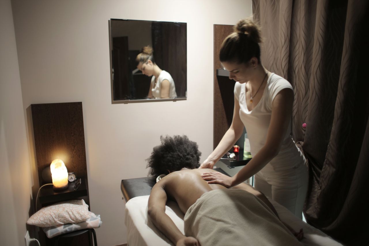 Professional masseuse in white uniform and doing massage of back of relaxed ethnic female client in modern salon